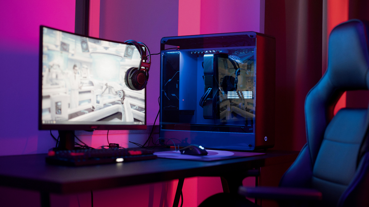 a gaming setup with a PC, monitor, headset, keyboard and mouse, a chair sits next to the desk. Purple lighting in various shades is in the background